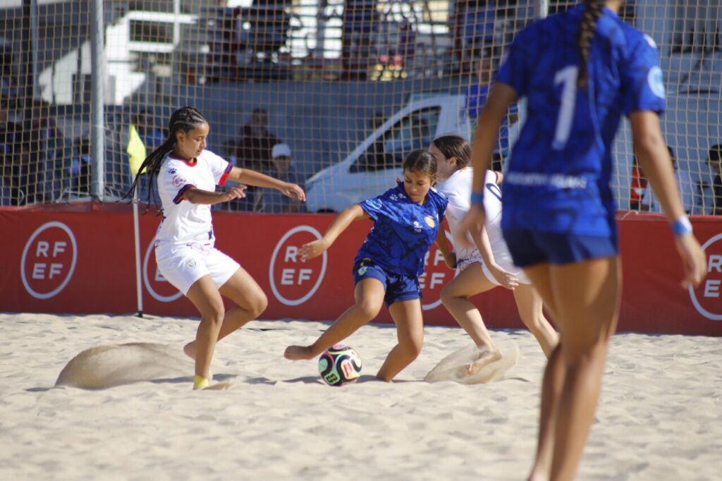 Girls' beach soccer match: a blue-uniform player kicks the ball while a white-uniform opponent tries to intercept on sandy field, with a red barrier in the background.