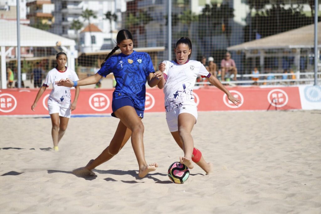 Two female beach soccer players chase the ball on a sandy court, one in blue and the other in white, with a stadium/boards in the background.