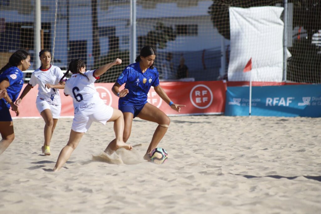 Women’s beach soccer match: a player in blue controls the ball while opponents in white chase on sandy ground near the goal banners.