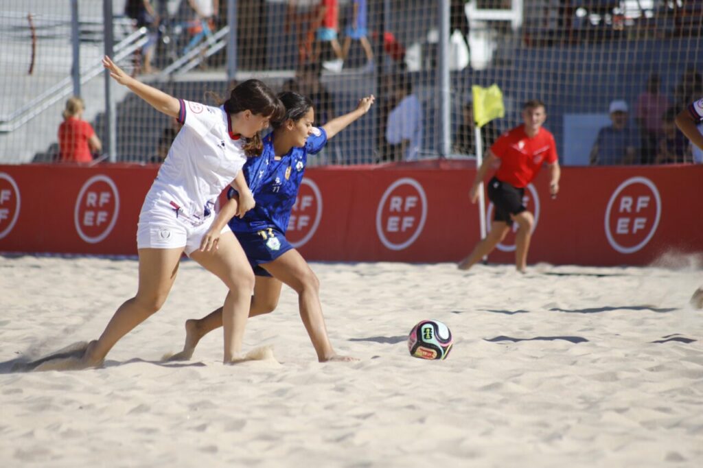 Two female players chase a colorful soccer ball on a sandy beach soccer field, white and blue uniforms, red advertising boards in the background