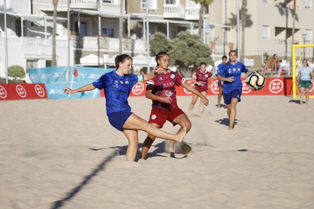 Women’s beach soccer match: a blue team player kicks the ball while a maroon teammate defends on a sandy court with buildings in the background in daylight.