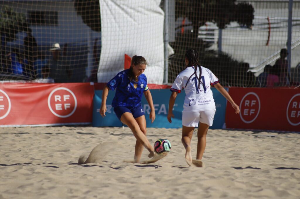 Two female beach soccer players chase a colorful ball on a sandy field, with red advertising banners in the background (RF EF).