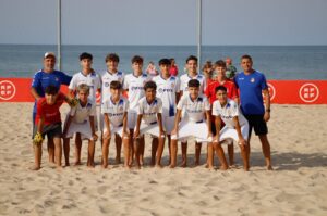 Youth soccer team in white uniforms posing on a sandy beach with two coaches in blue, ocean in the background and goal nets behind them.