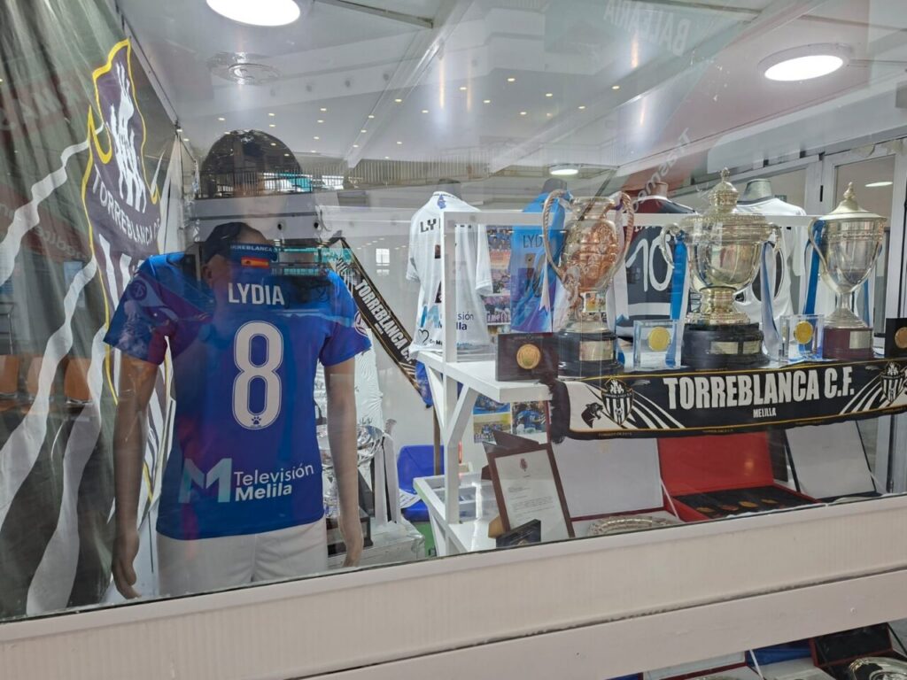 Display case of Torreblanca C.F. Melilla memorabilia featuring a blue Lydia 8 jersey, trophies, ribbons, and club banners.