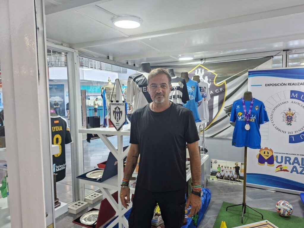 Man in a black T-shirt stands in a sports memorabilia booth with jerseys, trophies, and banners behind him.