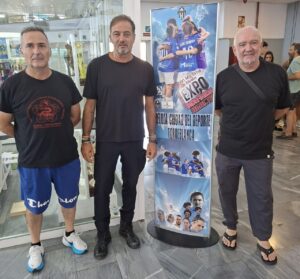 Three men in black shirts stand next to a tall blue poster advertising a sports expo with images of athletes.