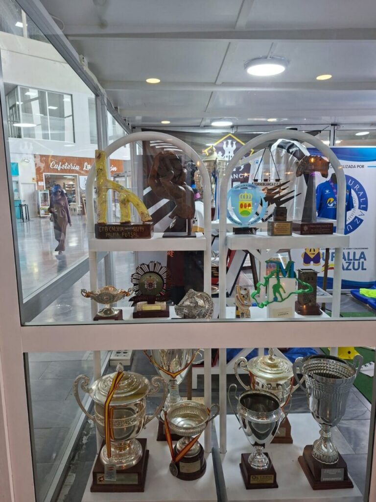 Shelf-filled display case with numerous trophies, cups, and plaques arranged on white shelves inside a glass cabinet, in a shopping mall corridor.