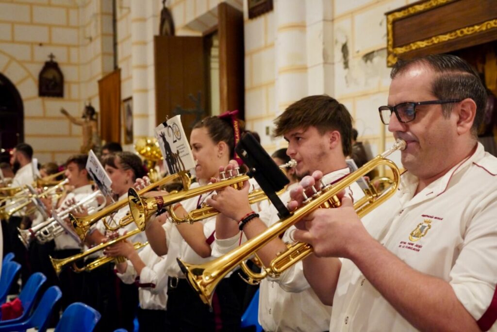 Brass band performing in a church, players in white shirts blowing trumpets as sheet music sits on stands in front of them