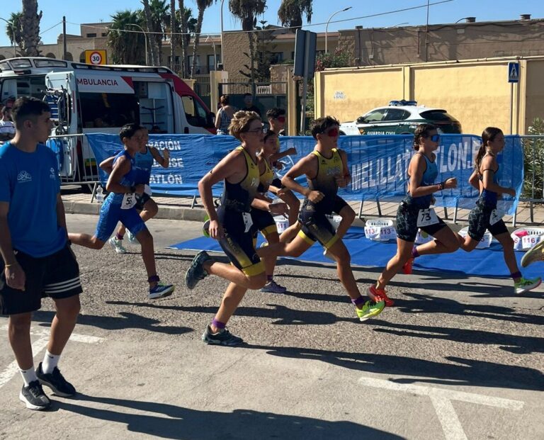 Group of young runners in a street race, wearing numbered bibs, with blue barriers and an ambulance in the background.