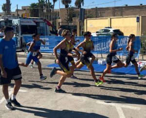 Group of young runners sprinting on a sunny street race, wearing numbered bibs with a blue barrier and ambulance in the background.
