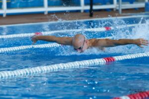 Swimmer wearing goggles performs butterfly stroke in a blue pool, arms extended and water splashing around them in a lane.