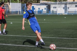 Young male soccer player in blue kit with a black training vest controls a soccer ball on a green artificial turf field, with a teammate in red nearby.