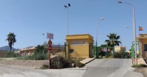 Gate and guardhouse at the entrance of a fenced facility, with a 20 km/h speed limit sign, palm trees, and a paved driveway.