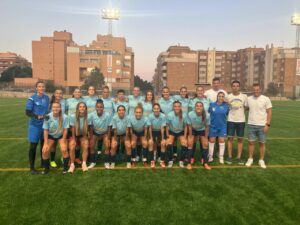 Group of female soccer players in light blue jerseys posing for a team photo on a field, with coaches at the ends and city buildings in the background.