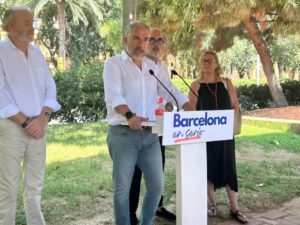 Man in a white shirt speaks at a podium with a Barcelona en Serio sign, outdoors under trees, with three colleagues nearby.