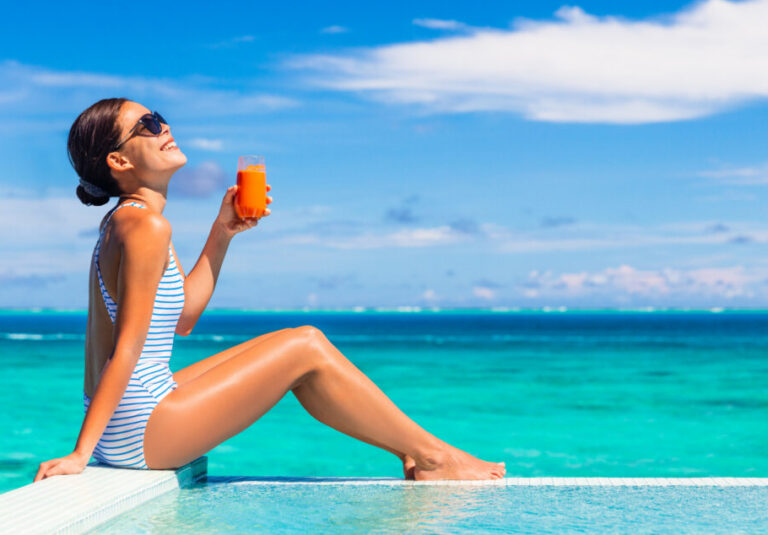 Woman in a striped swimsuit sits on the pool edge, holding an orange drink, with turquoise sea and blue sky behind her.”] ,