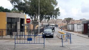Black car approaches a barrier checkpoint on a street, with blue metal barricades and a no-entry sign nearby.