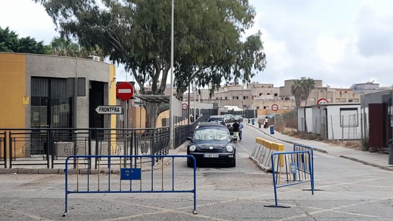 Black car approaches a barrier checkpoint on a street, with blue metal barricades and a no-entry sign nearby.