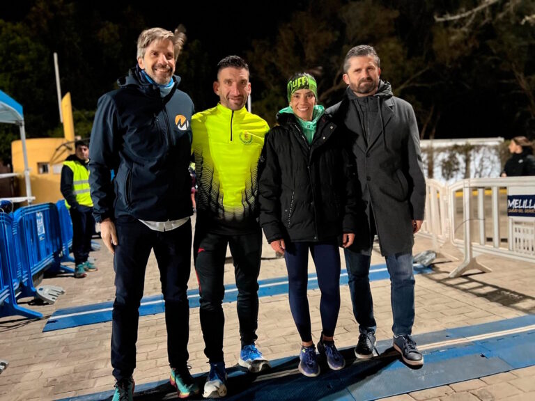 Four people pose together at night after a race, smiling, wearing athletic jackets and running shoes by a finish area barrier.
