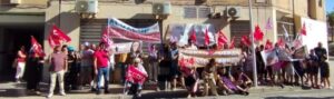 Crowd of protesters on a sidewalk with banners and red flags during a demonstration on a sunny day in front of buildings.