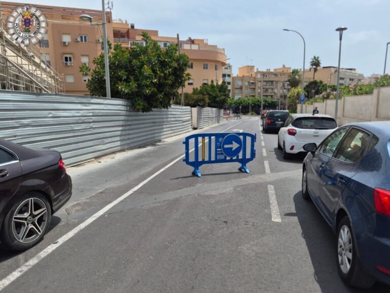 Blue portable barrier blocks a lane with a right-pointing arrow on a city street; cars lined up on both sides and construction fencing on the left.
