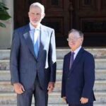 Two men in formal suits stand side by side on marble steps in front of a large wooden door.
