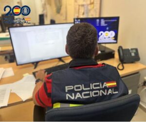 Policía Nacional officer sitting at a desk, facing two computer monitors, wearing a vest that reads POLICÍA NACIONAL with a Spanish flag patch.