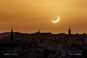 Silhouetted city skyline at sunset with a bright crescent sun in an orange sky; church spires and a tall antenna tower are visible along the horizon.