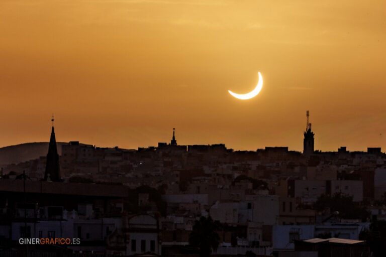 Silhouetted city skyline at sunset with a bright crescent sun in an orange sky; church spires and a tall antenna tower are visible along the horizon.