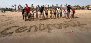 Grupo de niños y educadores en la playa con el nombre CentrosD2 en la arena