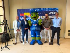 Four men pose with a green turtle mascot in blue sportswear at an indoor event, a camera on a tripod to the left and a banner behind them.