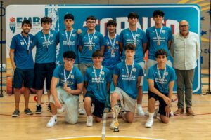 Group of young basketball players in matching blue jerseys posing for a team photo with medals around their necks and a trophy on the gym floor, coachs standing at ends, banner backdrop behind them.
