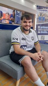 Smiling man in a white esports jersey sits on a gray sofa in a gaming venue, with banners in the background.