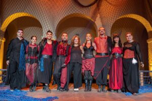 Group of performers in red and black costumes posing on stage with a net backdrop behind them, smiling at the camera.