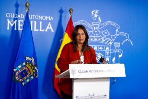 Woman in a red blazer speaks at a podium in front of a blue Melilla backdrop with flags and the city crest.