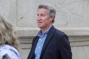 Man in a blue shirt and navy blazer walking outdoors beside a stone wall with a thoughtful expression.