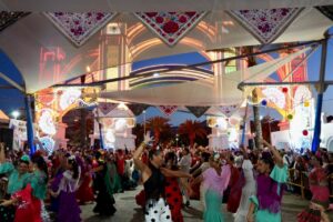 Celebration with dancers in colorful dresses under decorative tents and bright lights at dusk festival.