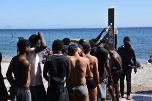 Group of shirtless men on a sunny beach forming a circle around a tall metal pillar as water is poured onto the crowd, with the sea in the background