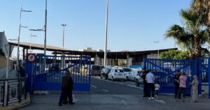 Blue gate blocks a roadway at a parking area; people stand nearby and cars line up behind the gate in clear weather.