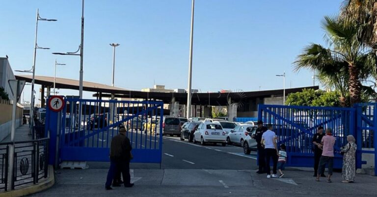 Blue gate blocks a roadway at a parking area; people stand nearby and cars line up behind the gate in clear weather.