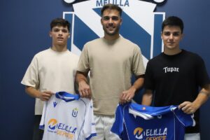 Three young men stand in front of a blue Melilla crest wall, each holding a blue-and-white team jersey with 'Melilla Deporte' logos, smiling at the camera.