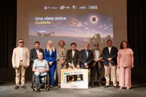 Group of ten people on a stage posing for a photo, one person in a wheelchair, with a large framed card in front displaying a solar eclipse image and logos behind them.