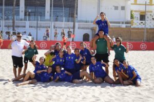 Group of female soccer players in blue uniforms pose on sandy beach with a teammate on someone's shoulders, celebrating together.