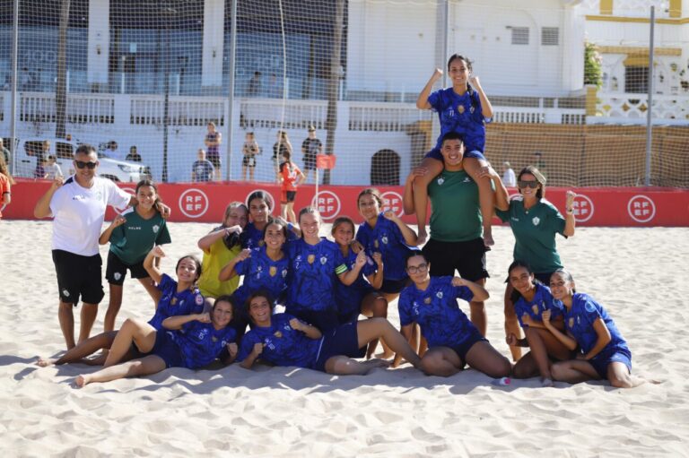 Group of female soccer players in blue uniforms pose on sandy beach with a teammate on someone's shoulders, celebrating together.