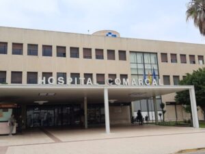 Facade of a hospital with large sign 'HOSPITAL COMARCAL' and a covered entrance area with glass doors and flags nearby, daylight.