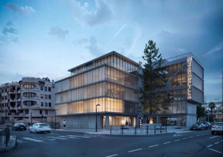 Modern glass-faced office building at a street corner, with warm interior lights and a tall tree nearby.