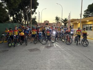Group of cyclists in colorful jerseys posing with bikes on a cobblestone street at sunset.