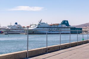 Harbor scene with large white passenger ferries docked beyond a chain-link fence under a clear blue sky
