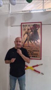 Man in a black T-shirt holding a microphone, standing beside a framed bullfighting poster on a white wall.