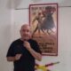 Man in a black T-shirt holding a microphone, standing beside a framed bullfighting poster on a white wall.
